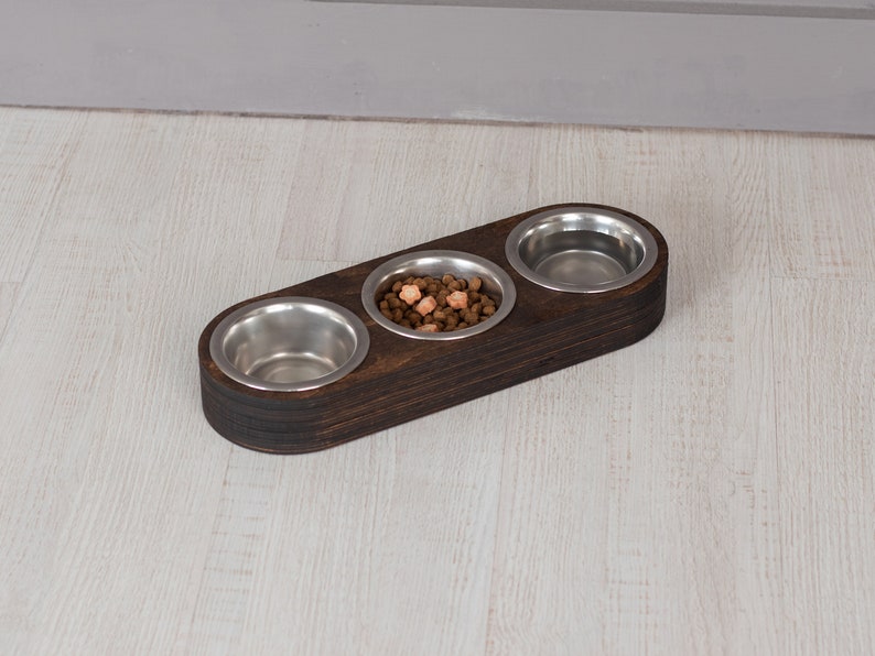 May include: A dark brown wooden pet food bowl with three stainless steel bowls. The center bowl has dry pet food in it.