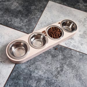 May include: A wooden pet food bowl holder with four stainless steel bowls. One bowl is filled with brown pet food.
