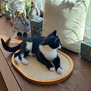 May include: A black and white cat laying on a round, beige scratching post with a wooden frame. The cat is looking to the right of the image. The scratching post is on a wooden surface.