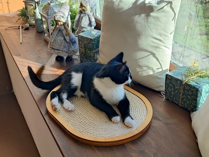 May include: A black and white cat laying on a round, beige scratching post with a wooden frame. The cat is looking to the right of the image. The scratching post is on a wooden surface.