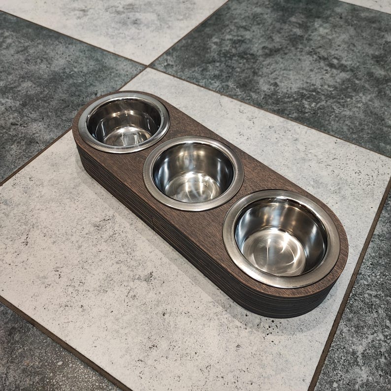 May include: A wooden pet food bowl holder with three stainless steel bowls. The holder is dark brown and has a rounded rectangular shape.