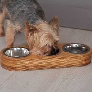 May include: A small dog eats from a wooden elevated dog bowl with three stainless steel bowls. The bowl is made of light brown wood and has a natural wood grain.