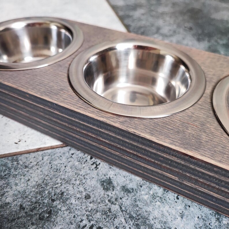 May include: A wooden pet food bowl stand with three stainless steel bowls. The stand is made of dark wood with a natural wood grain finish.