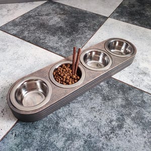 May include: A wooden pet food bowl holder with three stainless steel bowls. The holder has a dark gray finish and is designed to hold food and water for pets. The holder is shown on a gray and white tiled floor.