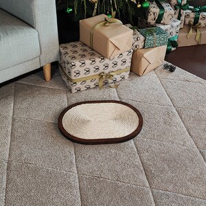 May include: A light brown oval-shaped cat scratching post with a woven rope surface. The scratching post is on a light gray carpet with a diamond pattern.