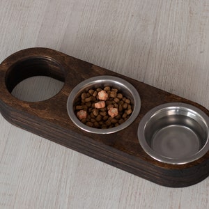 May include: A dark brown wooden pet food bowl stand with two stainless steel bowls. One bowl is filled with brown kibble and orange treats.