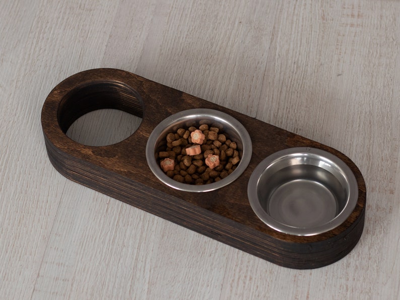 May include: A dark brown wooden pet food bowl stand with two stainless steel bowls. One bowl is filled with brown kibble and orange treats.