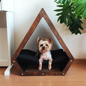 May include: A small, brown wooden dog bed shaped like a triangle with a black cushion inside. A small, brown and white Yorkshire Terrier dog is sitting inside the bed looking at the camera.