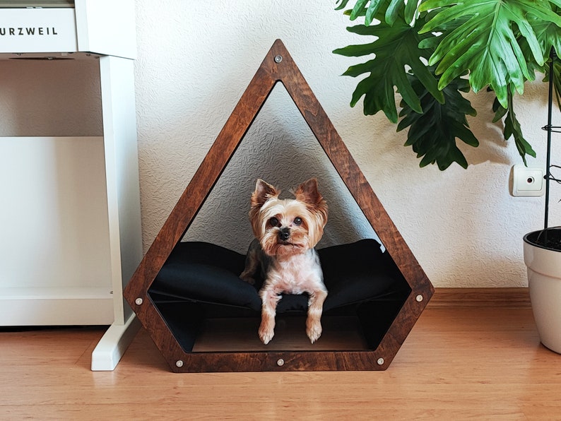 May include: A small, brown wooden dog bed shaped like a triangle with a black cushion inside. A small, brown and white Yorkshire Terrier dog is sitting inside the bed looking at the camera.