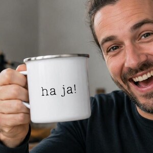 Può includere: Una tazza in smalto bianco con bordo color argento che presenta il testo "ha ja!" stampato in un semplice carattere nero in stile macchina da scrivere sulla parte anteriore. La tazza ha un manico robusto e una finitura liscia e lucida.