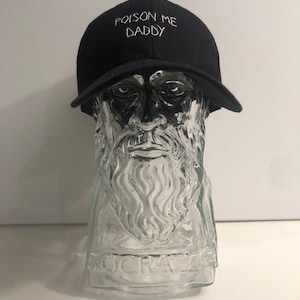 May include: A black baseball cap with a white embroidered phrase "POISON ME DADDY" on the front.