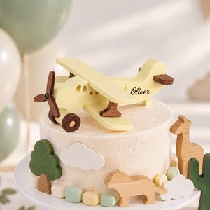 Pode incluir: Um topo de bolo em forma de avião de madeira amarelo-claro com o nome Oliver impresso na lateral, apresentando hélice e rodas de madeira castanho-escura. O bolo é decorado com recortes de madeira de uma girafa, um leão, uma árvore, um cacto e nuvens.