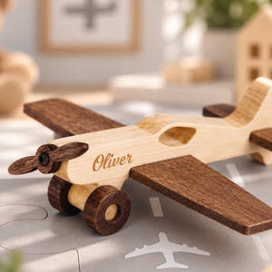 Pode incluir: Um avião de brinquedo de madeira com corpo de cor clara e asas, hélice e rodas de cor escura. O nome Oliver está gravado em letra cursiva na lateral da fuselagem. O brinquedo está posicionado sobre uma superfície cinza com gráficos de aviões brancos.