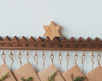 May include: A wooden advent calendar with 25 numbered envelopes hanging from a wooden bar. Each envelope is decorated with a small sprig of greenery and a red berry. The numbers 9 through 25 are visible on the bar.
