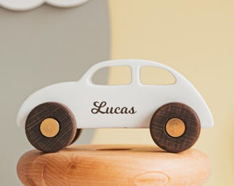 May include: A white wooden toy car with dark brown wheels and gold centers, featuring the name Lucas printed in black script on the side. The car is balanced on top of two smooth, rounded wooden rings, one light brown and one white in color.