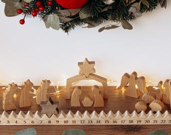 May include: A festive Christmas display featuring a wooden advent calendar with numbers 1-24, wooden nativity figures, and a wreath with red and white baubles. The scene is decorated with fairy lights and red berries.