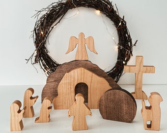 May include: Wooden figurines depicting the Easter story, including an angel, a tomb, and figures kneeling in prayer. The figures are arranged on a wooden base with a brown stone tomb and a wooden angel above it. The figures are all made of natural wood and have a rustic, handmade look.