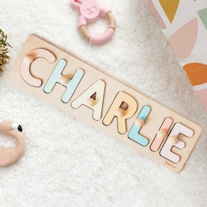 May include: Wooden name puzzle spelling "CHARLIE" in pastel colors. Includes a pink bunny teether and a flamingo-shaped teether. A decorative pillow with abstract shapes and a small plant are also in the image. A baby toy.