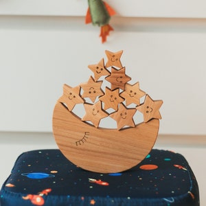 May include: A wooden toy featuring a crescent moon base with a smiling face and stacked star shapes. The toy is made of light-colored wood and sits on a navy blue ottoman with a space-themed pattern. Felt stars and a rocket hang in the background.