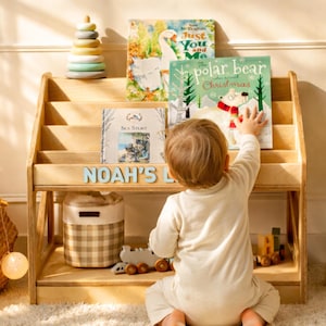 Puede incluir: Una estantería de madera clara de tres niveles, con el nombre NOAH'S en letras azul claro. El estante contiene varios libros infantiles, incluido The Polar Bear Who Saved Christmas, y un juguete de anillos apilables en colores pastel.