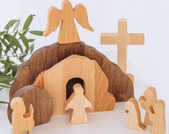 May include: Wooden toy set depicting the Easter story. The set includes a tomb, an angel, Jesus, and three figures. The tomb is made of brown wood and has a light brown door. The angel is standing on top of the tomb with its wings spread. Jesus is standing in front of the tomb with his arms outstretched. The three figures are kneeling in front of the tomb.