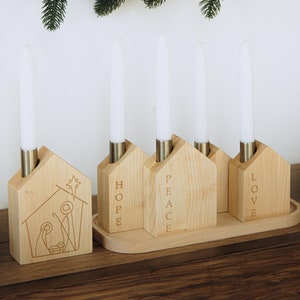 Könnte beinhalten: Kerzenhalter aus Holz mit vier Kerzenplätzen. Jeder Platz hat ein hausförmiges Design mit den Worten "Hope", "Peace", "Love" und einer Weihnachtskrippe.