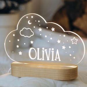 May include: A white acrylic night light shaped like a cloud with stars and a crescent moon. The name "Olivia" is engraved on the cloud. The light sits on a wooden base.