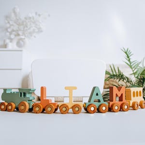 Könnte beinhalten: Ein hölzernes Spielzeugeisenbahn-Set mit einzelnen Waggons, die den Namen LIAM bilden. Der Zug besteht aus einer salbeigrünen Lokomotive, gefolgt von orangefarbenen, gelben, salbeigrünen und orangefarbenen Buchstabenwaggons und einem gelben Waggon.