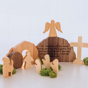 Può includere: Scena pasquale in legno con un angelo, una croce e delle figure. La scena comprende una tomba e una roccia con testo inciso da Giovanni 3:16. Accenti floreali verdi sono posti attorno alle figure. Le figure sono in legno chiaro.