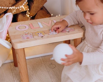 May include: A wooden children's stool with the name "OLIVIA" in pink letters. The stool features princess-themed illustrations and is supported by four wooden legs. The child is playing with a white ball.
