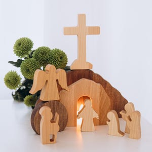 Può includere: Una scena pasquale in legno con una croce, un angelo e delle figure vicino a una tomba. La scena è realizzata in legno chiaro e include una piccola candela accesa all'interno della tomba. Fiori verdi sullo sfondo.