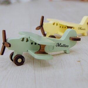 Puede incluir: Dos aviones de juguete de madera, uno de color verde claro y otro amarillo, con hélices, ruedas y detalles de cola en color marrón oscuro. El avión verde tiene el nombre Matteo impreso en el lateral y el amarillo tiene el nombre Oliver.