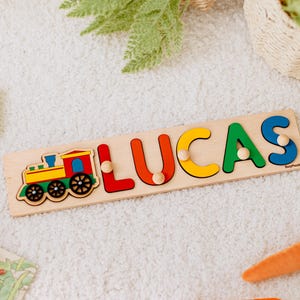 Peut inclure: Puzzle en bois avec le nom "LUCAS" composé de lettres colorées et d'un dessin de train. Les lettres sont rouges, orange, jaunes, vertes et bleues. Le train est jaune, rouge, vert et bleu. Le puzzle est sur une surface claire avec des carottes et de la verdure.