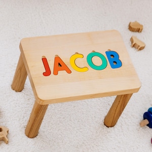 May include: A wooden children's stool with the name "JACOB" spelled out in colourful puzzle letters. The stool has four wooden legs and is surrounded by wooden toys, including a stackable ring toy and animal shapes. The stool is on a white rug.