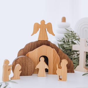 May include: Wooden Easter scene featuring a tomb, angel, cross, and figures. The tomb is made of light and dark brown wood. The angel has outstretched wings. The figures are in various poses. The scene is set on a white surface with greenery.