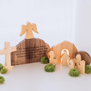 Può includere: Scena pasquale in legno con croce, angelo e figure. La scena include un arco in legno e un cartello con il testo "For God so loved the world..." e Giovanni 3:16. Accenti floreali verdi sono posizionati intorno.