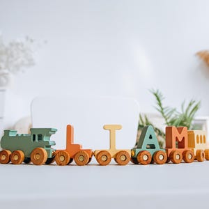 May include: A wooden toy train set featuring a green locomotive followed by individual train cars shaped like the letters L, I, A, and M, finished with a yellow caboose. Each car has natural wood wheels with gold-colored centers.