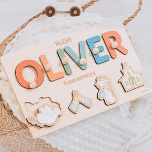 May include: Wooden name puzzle with the name "OLIVER" in colorful letters. The puzzle includes additional wooden shapes of a dove, praying hands, an angel, and a church. A small wooden car sits on top of a woven basket.