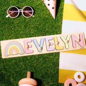 May include: A wooden puzzle spelling "EVELYN" in pastel colors, with a rainbow design. The puzzle is on a green surface with sunglasses, a party hat, and other toys. The letters have wooden pegs for easy handling.