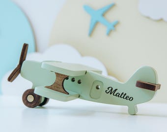 May include: A light green wooden toy airplane featuring dark brown accents on the propeller, wheels, and wing supports. The side of the airplane is personalized with the name Matteo in a black script font, showcasing a classic biplane design.