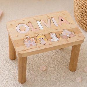 May include: A small wooden step stool with the name "OLIVIA" spelled out in pink letters. The stool features princess-themed puzzle pieces and is supported by four wooden legs. The stool measures approximately 10 inches long.