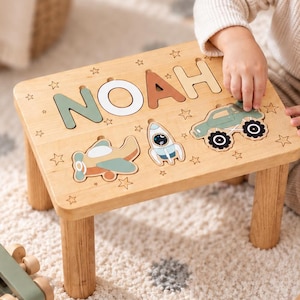 May include: A rectangular wooden stool featuring a puzzle top with the name NOAH in large letters, accompanied by cutouts of an aeroplane, a rocket ship, and a monster truck. The surface is decorated with small star engravings throughout the wood.