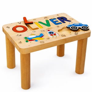 May include: A wooden children's table with the name "OLIVER" in colourful, raised letters. The table has a car, aeroplane, and rocket ship puzzle pieces. The table is made of light-coloured wood and has four sturdy legs.