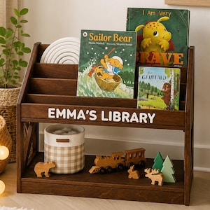 May include: A dark wood children's bookshelf with three slanted shelves for books and a bottom storage shelf. The front features the white text EMMA'S LIBRARY. It holds three children's books, a beige checkered fabric bin, and wooden animal toys.