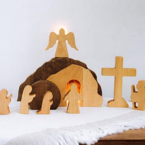 Può includere: Scena pasquale in legno con un angelo, una croce, un sepolcro e delle figure. Il set include un angelo colorato chiaro in cima a una collina marrone scuro, una croce e diverse figure. Il sepolcro ha una luce all'interno e le figure sono in varie pose.
