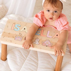 May include: A light-colored wooden puzzle stool featuring the name ISABELLA in pastel-colored cutout letters. The surface includes engraved illustrations of a castle, a unicorn, and a carriage. The stool has four cylindrical wooden legs.