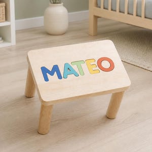 May include: A light-coloured wooden stool with four sturdy legs and a flat rectangular top. The surface of the stool features the name MATEO inlaid in large, colourful, individual wooden letters in blue, light blue, green, yellow, and orange.