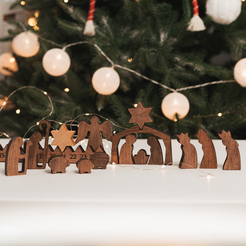 May include: Wooden nativity scene with figures of Mary, Joseph, baby Jesus, and the three wise men. The figures are arranged in front of a wooden stable with a star on top. The scene is set against a backdrop of a Christmas tree with lights.