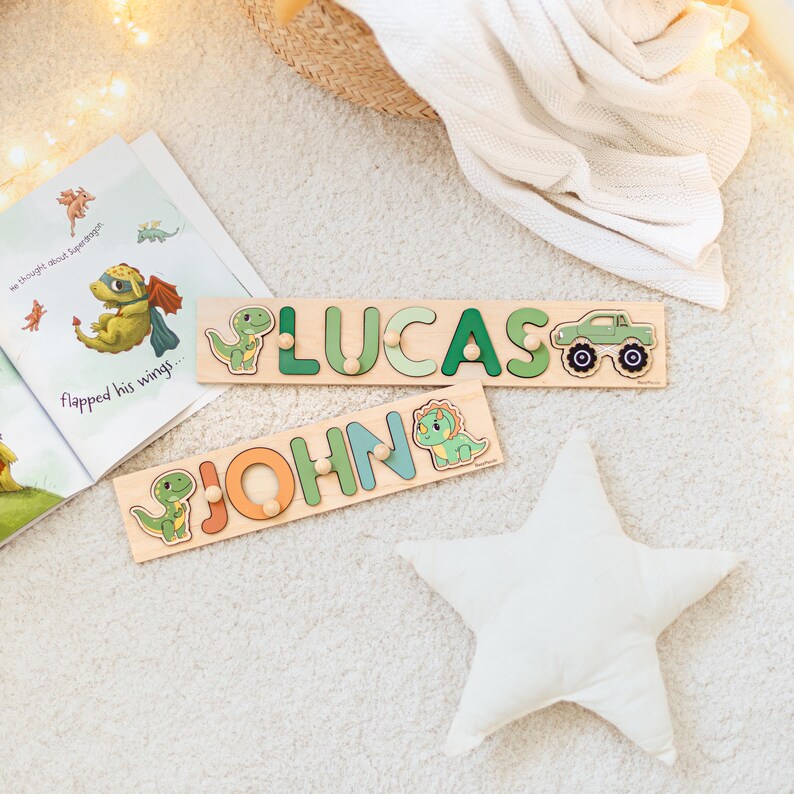 May include: Wooden name puzzles with dinosaur and truck designs. One puzzle spells "LUCAS" in green letters, and the other spells "JOHN" in orange and blue letters. A children's book with a dragon illustration is open nearby.