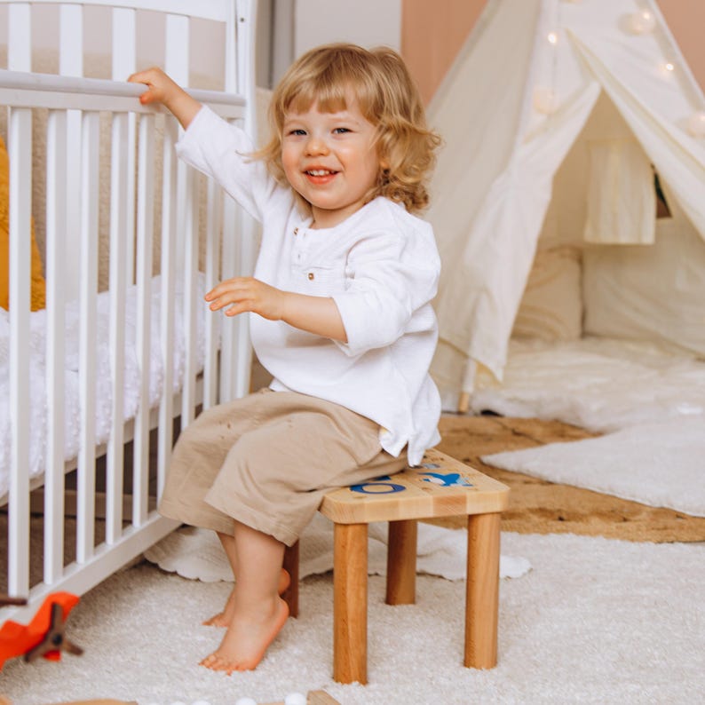 May include: A small wooden stool with four cylindrical legs and a flat rectangular top featuring blue and black graphic illustrations of a whale and other sea creatures, positioned on a white rug near a white crib and a white fabric play tent.